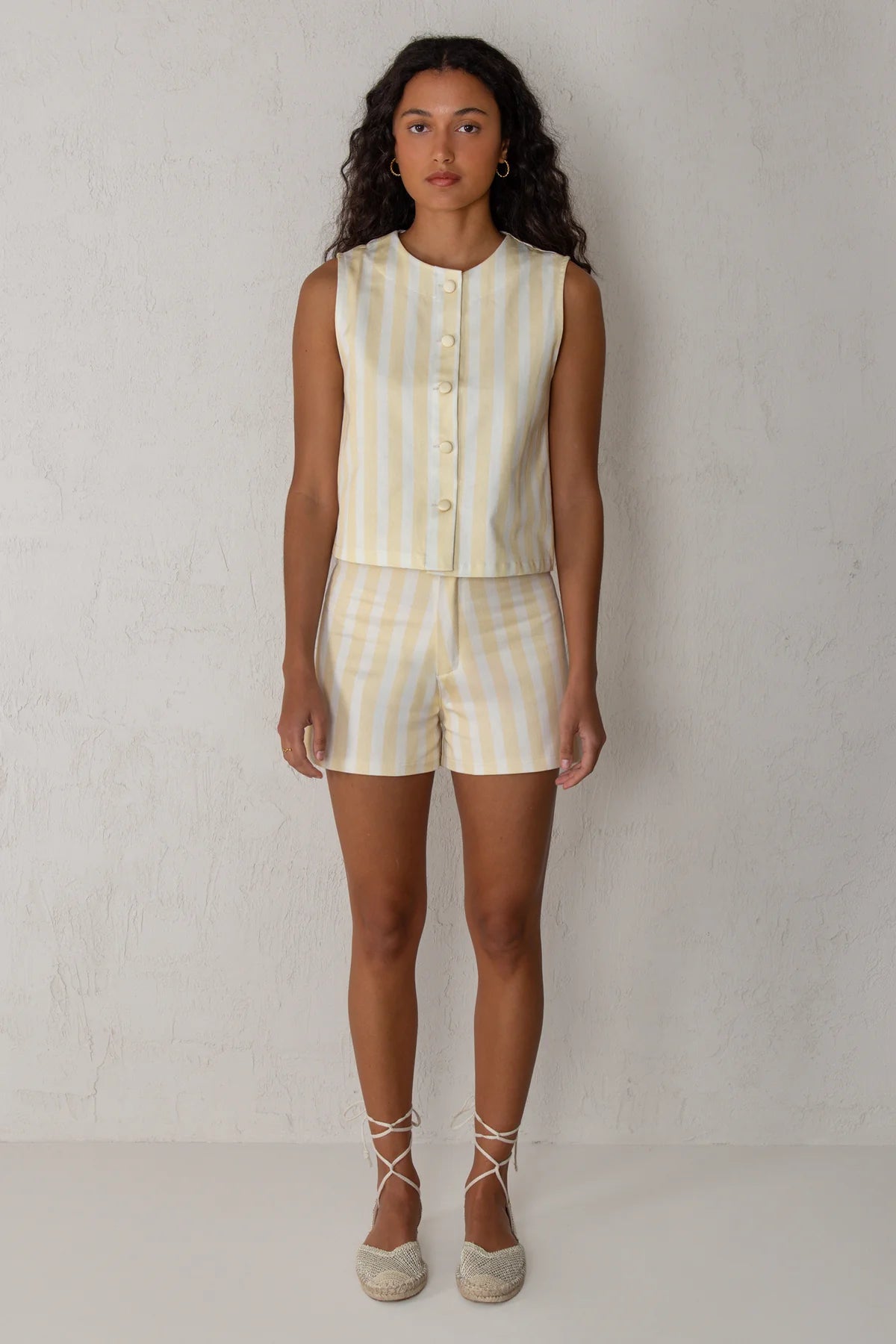 A woman stands against a light textured wall wearing a sleeveless cream and pale yellow striped vest and matching striped shorts, paired with beige lace-up espadrille flats.